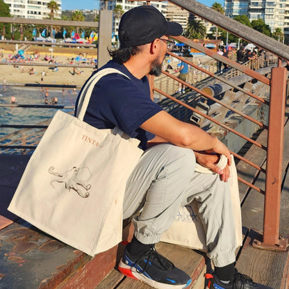 Persona sentada junto al mar llevando bolsa de tela TINTAmar con diseño de pulpo dibujado a mano