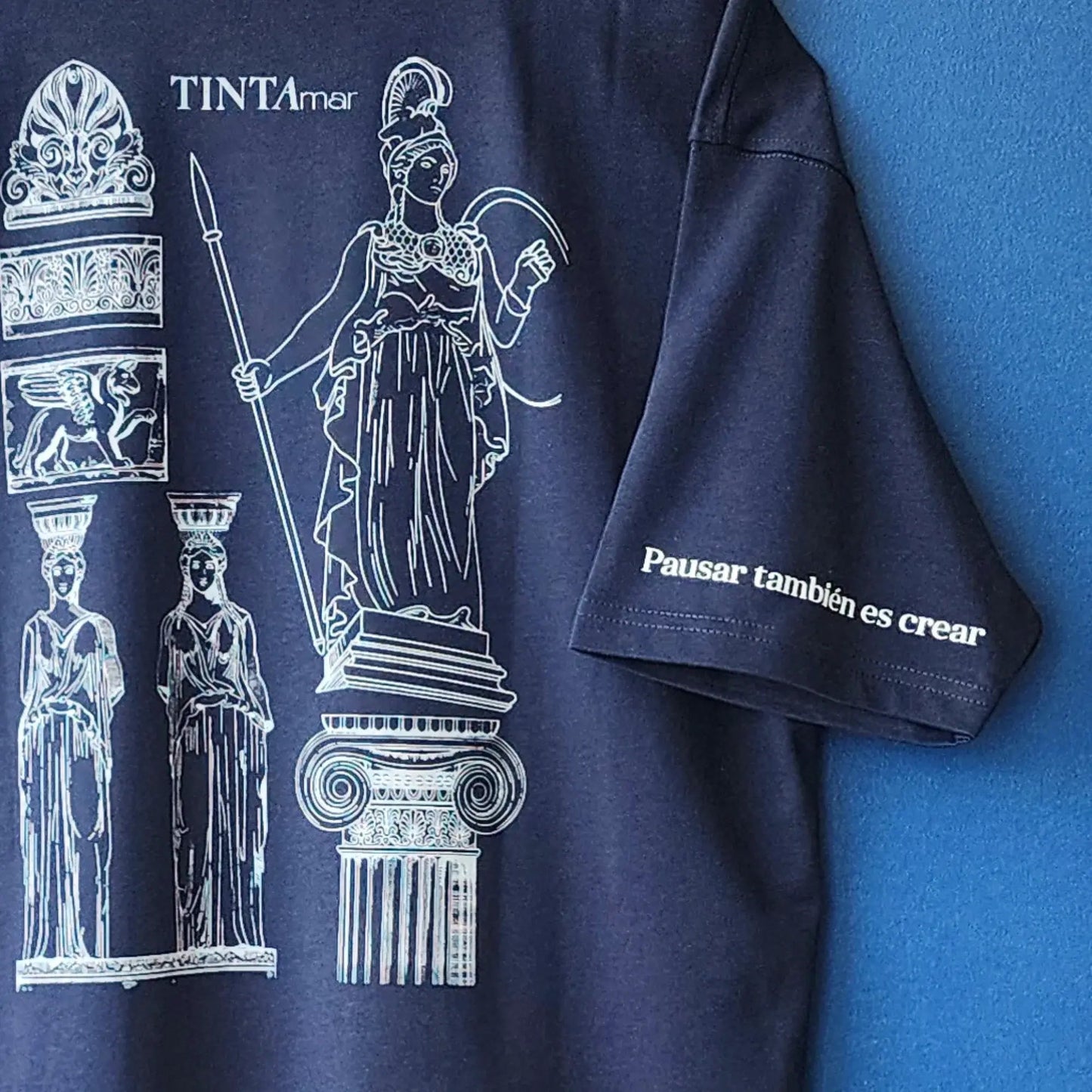 Detalle de polera azul con diseño de Atenea y cariátides, frase “Pausar también es crear” en la manga